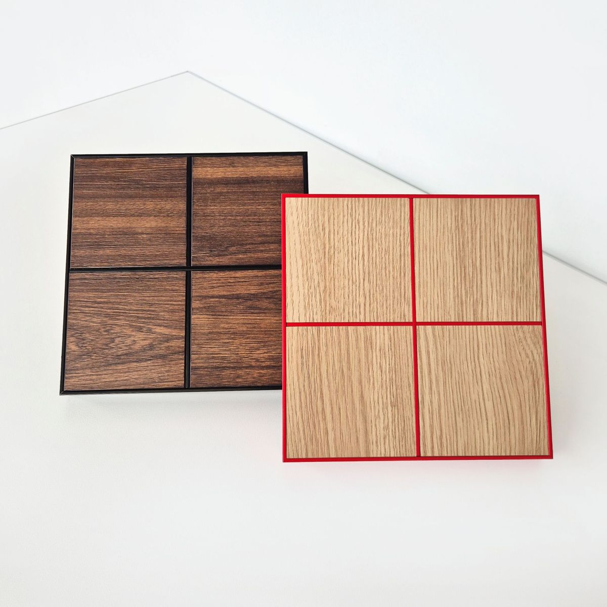 Two square sample boards showing woodgrain finishes are displayed on a clean white background. The left sample has a dark brown woodgrain pattern with black powder-coated Stickman joinery, and the right sample has a light beige woodgrain pattern with red Stickman powder-coated joinery.