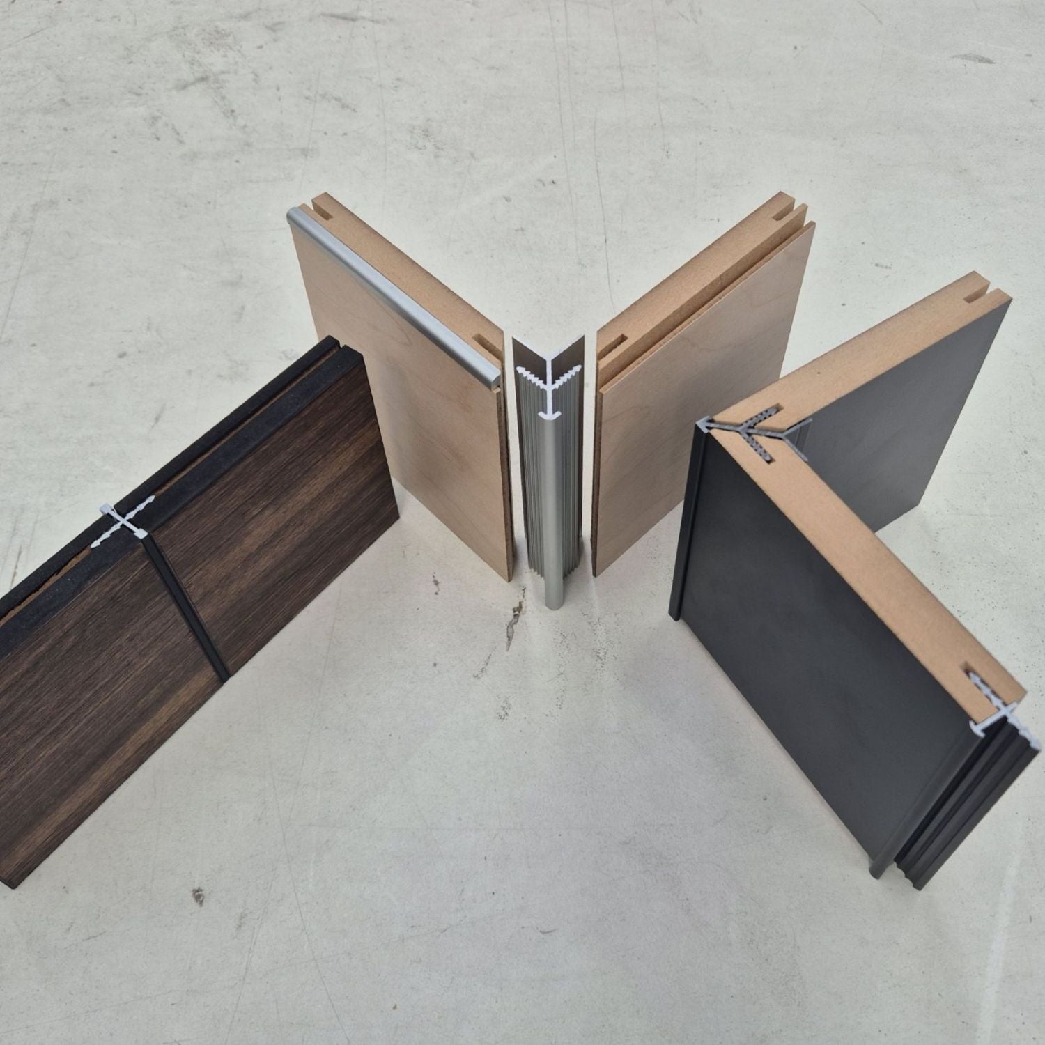 Four sample panels joined with Stickman profiles arranged on a light concrete floor, showing different finish options: two dark woodgrain pieces, two black pieces, and two natural wood pieces.